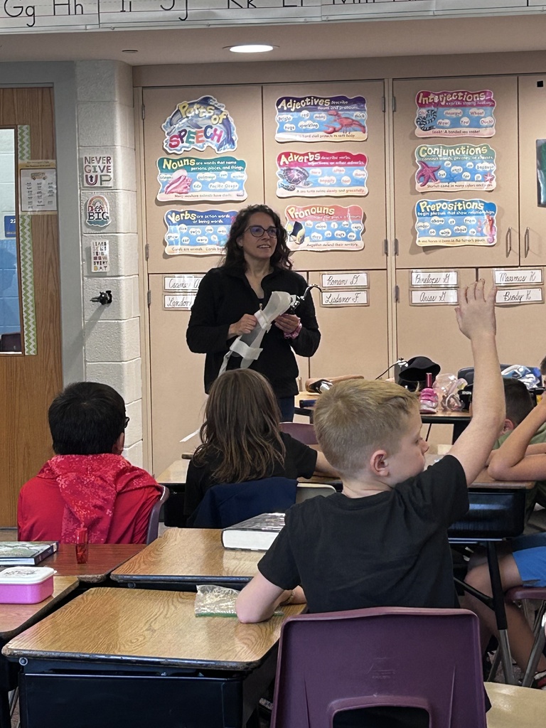 A student examines a prosthetic leg with a curved metal attachment while a presenter holds it. Additional prosthetic devices are displayed on a table in the classroom.