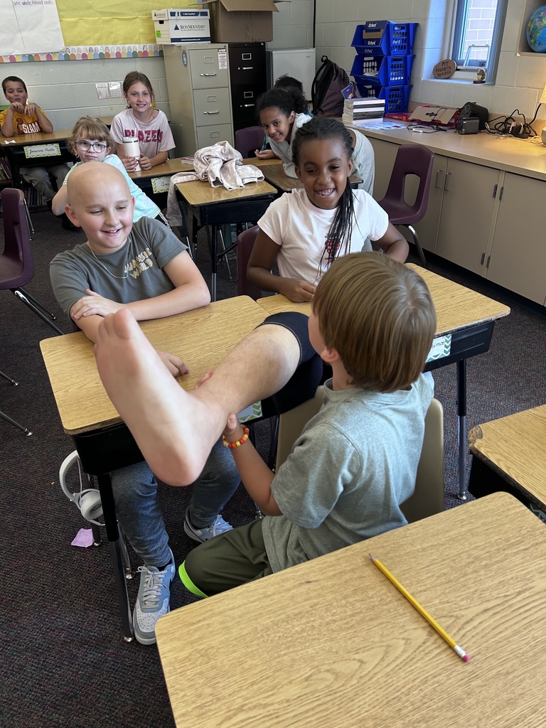 A student, seated at his desk, holds a prosthetic leg while his classmates smile.