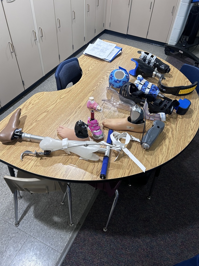 A variety of prostheses and orthoses are displayed on a classroom table, including prosthetic legs, feet, hands, and supportive braces in different sizes, colors, and designs.