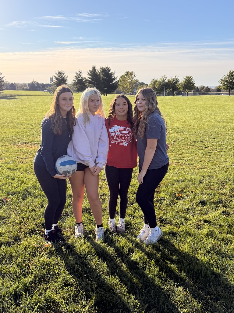 Four students outside on a field posing with a volleyball