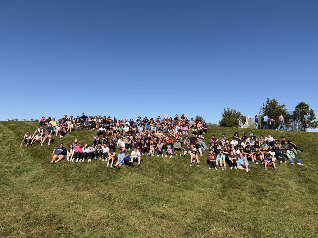 Large group of students sitting outside on a grassy hill.