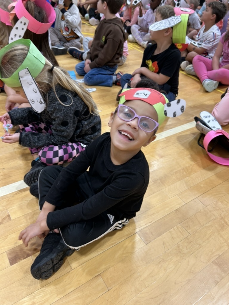 Picture of a student smiling at the camera while his classmates are seated near him in cross legged fashion.