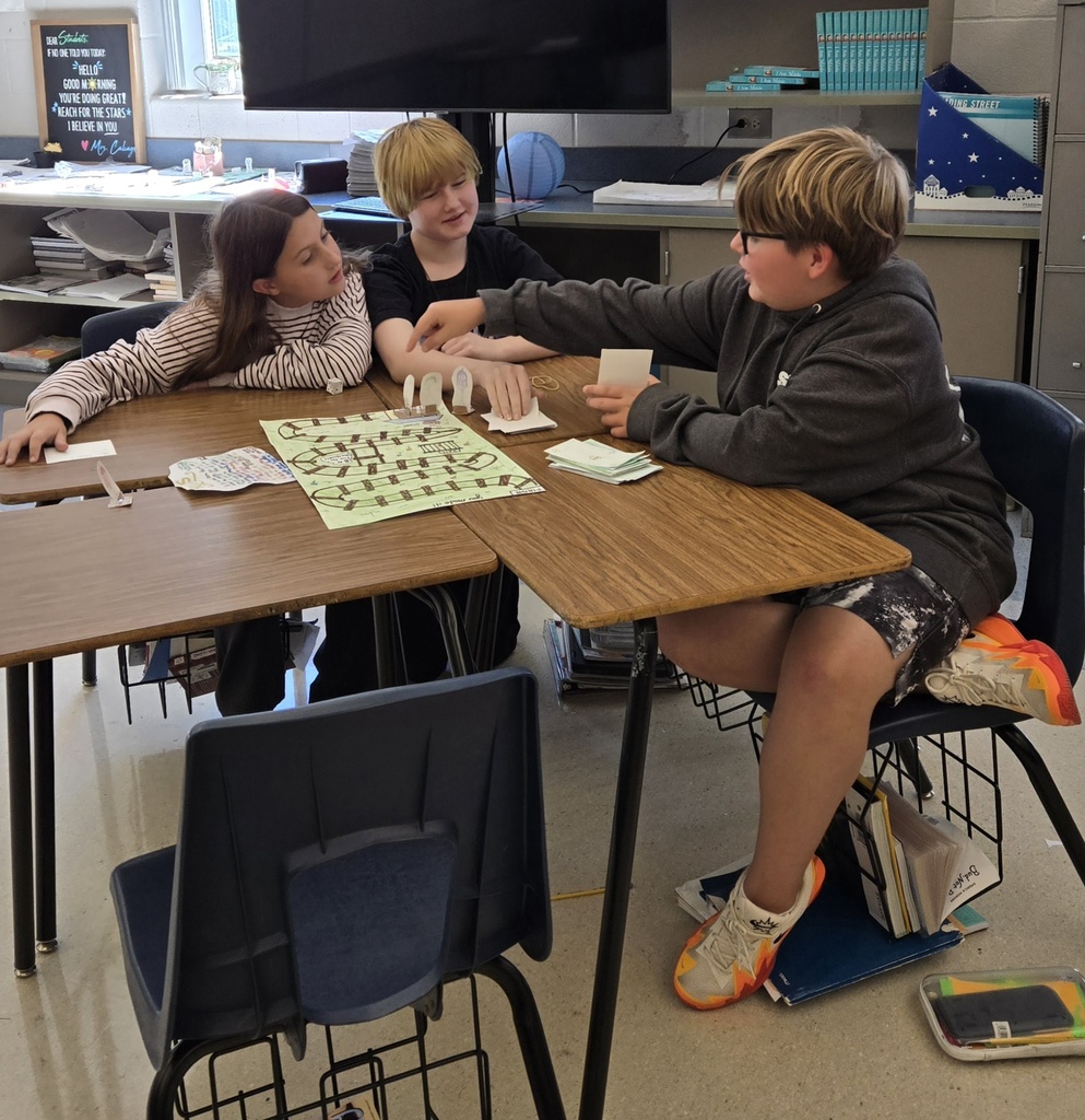 Three students gather around a table to examine and play a handmade board game, moving game pieces along a colorful path while referring to cards spread across the table.