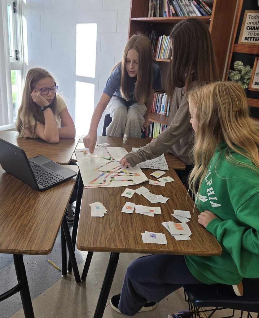Four students gather around a table to examine and play a handmade board game, moving game pieces along a colorful path while referring to cards spread across the table.