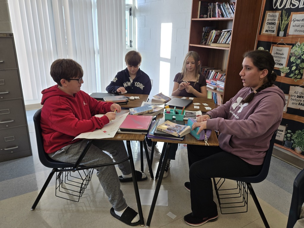 Four students sit around a classroom table, discussing and playing a handmade board game with paper game pieces and cards spread across the table.