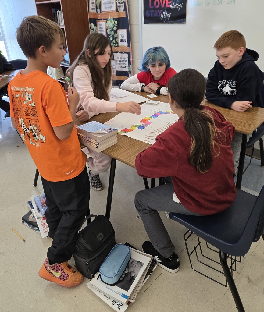 Five students work together around a classroom table, playing a handmade board game with a colorful path and numbered spaces.