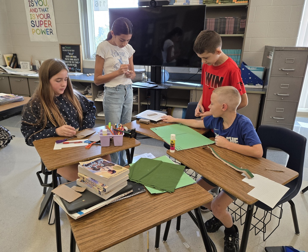 Four students collaborate on a board game project, cutting and arranging green paper and cardboard to create their game while supplies and books sit on nearby desks.