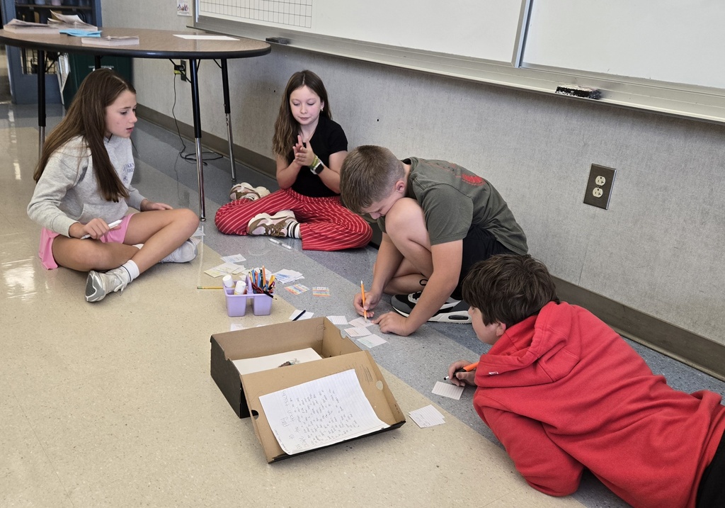 Four sixth-grade students sit on the classroom floor, working together to write game cards and develop a board game inspired by their reading.