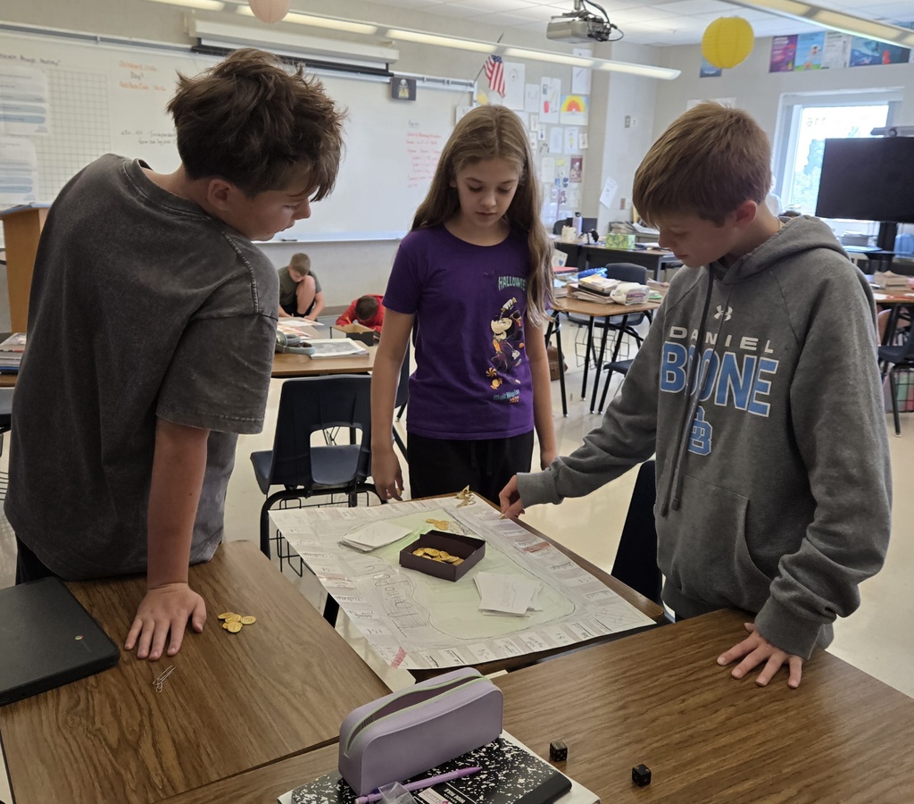 Three students stand around a desk to play a student-created board game, examining the game board and using small tokens as they follow the path.