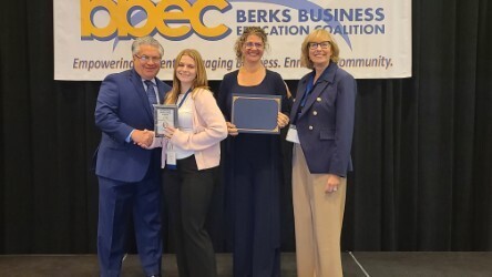 Four people stand on a stage in front of a BBEC Berks Business Education Coalition banner. Student is shaking hands with a man while holding a wooden plaque. One woman is holding a blue and gold certificate cover. All are smiling.
