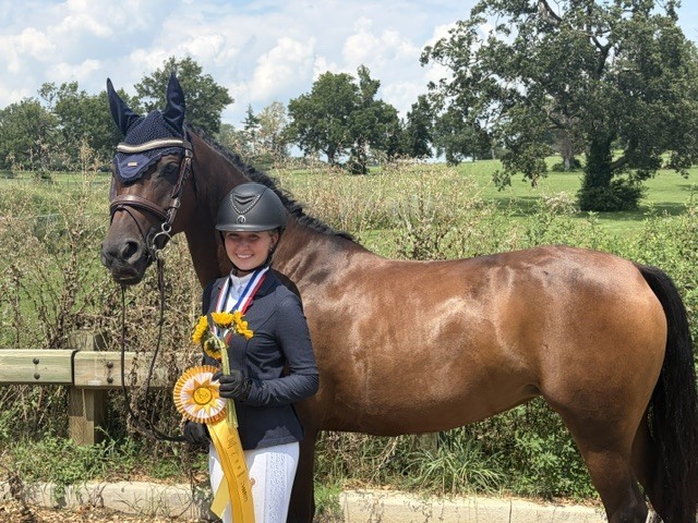 Student standing in front of a horse wearing riding gear and holding a yellow ribbon and flowers.