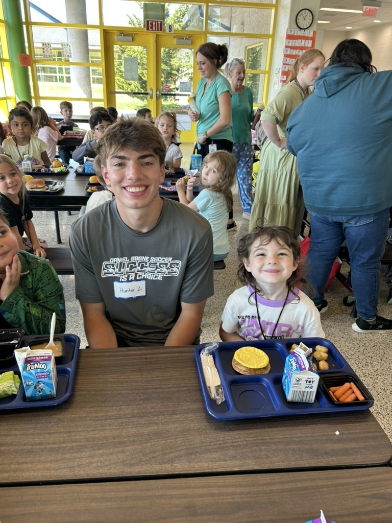 Primary Center student sits at a lunch table with high school student. Blue tray with primary students lunch sits on a table in front of them.