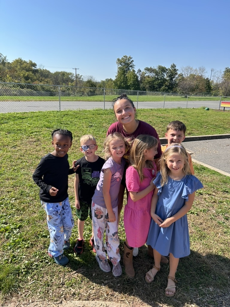 High school student standing with six primary center students in the grassy area of the playground. All are smiling for the camera.