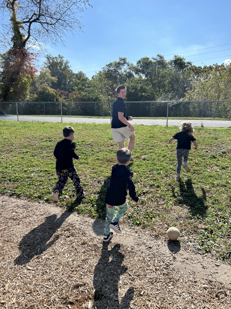 High school student with three primary center students kicking a ball on a grassy field of the playground.