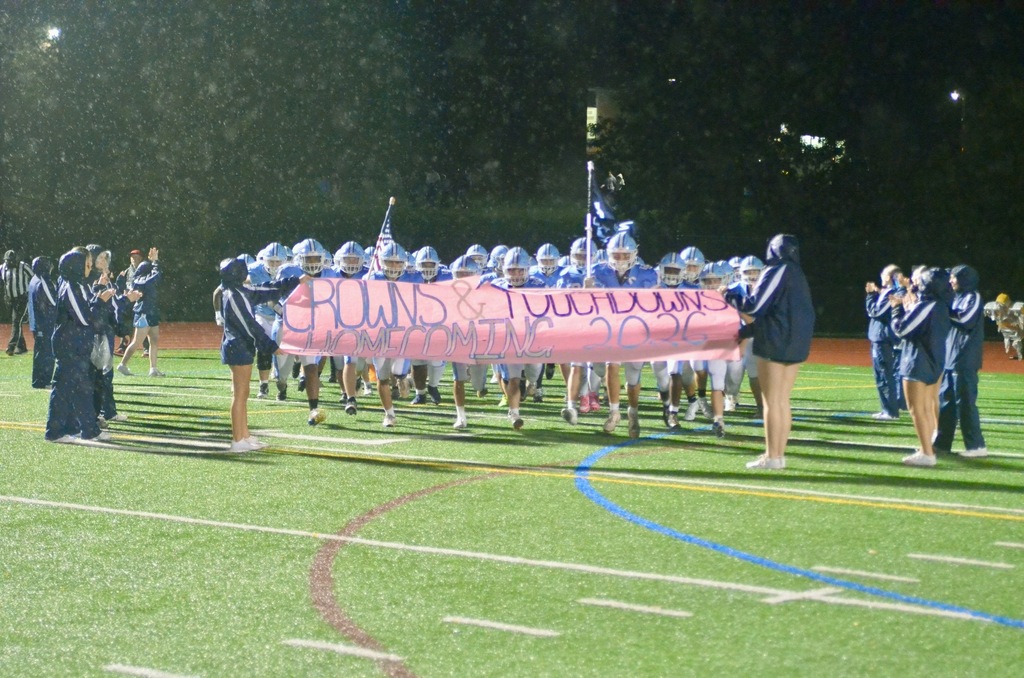 Football team prepares to run through a banner that reads Crowns & Touchdowns Homecoming 2026