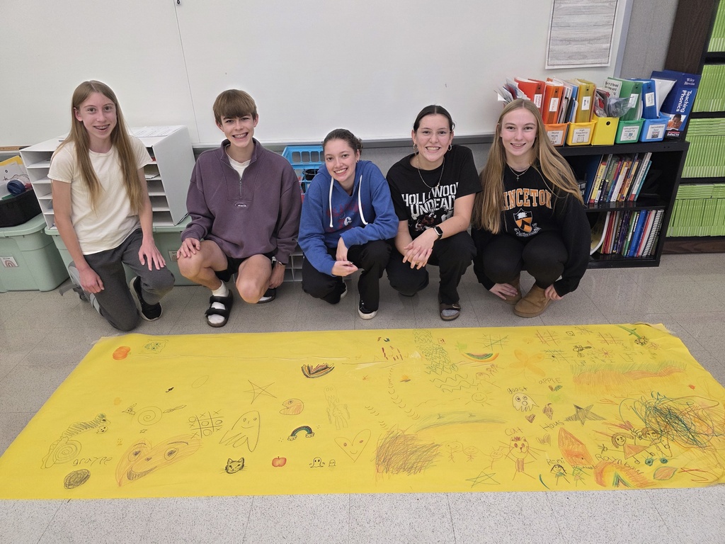 Five students kneeling in front of a large yellow piece of paper with drawings on it.