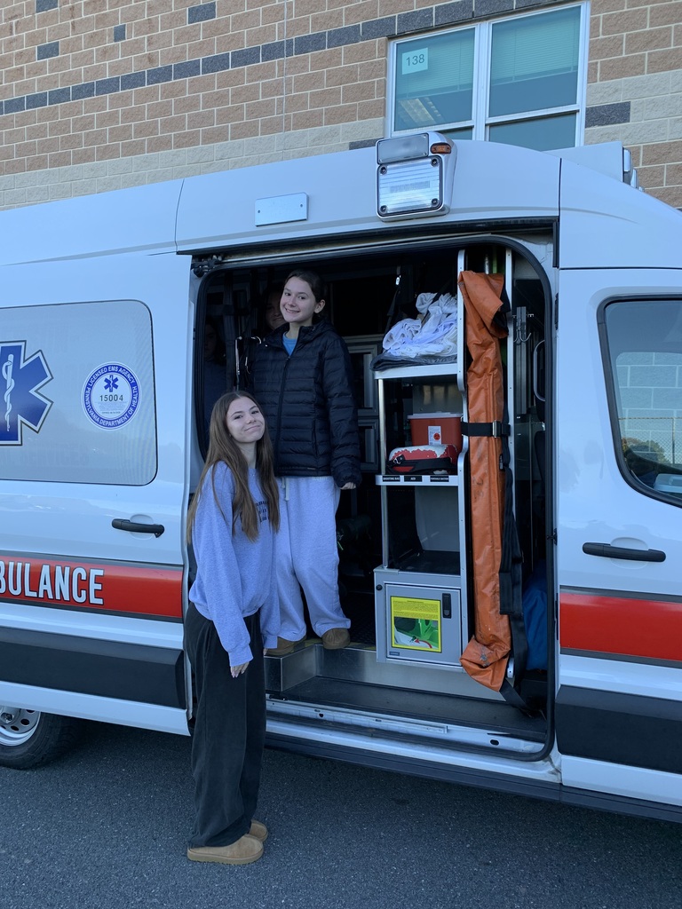 Two students - on standing inside an ambulance and one standing outside of the ambulance smile for the camera.