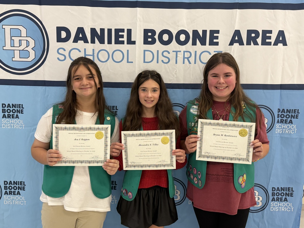 Three Girl Scouts wearing their green Girl Scout vests hold their certificates and smile in front of a Daniel Boone Area School District banner