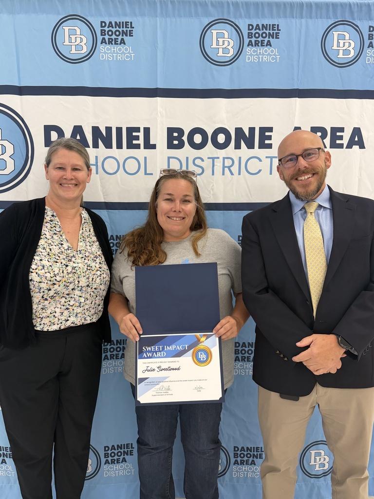 Melanie Hefter, Julie Sweetwood and Tom Voelker stand in front of a DBASD step and repeat banner. Julie is holding her Sweet Impact Award certificate. All are smiling.