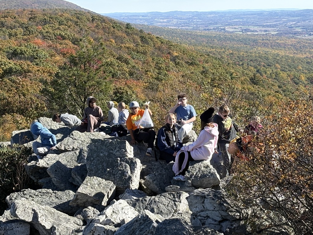 Sixth-grade students gather on a rocky summit at Hawk Mountain, surrounded by colorful fall foliage and scenic mountain views.