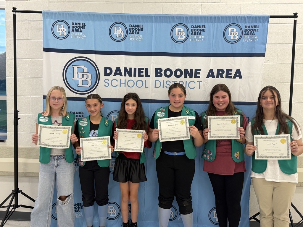 Six Girl Scouts wearing their green Girl Scout vests hold their certificates and smile in front of a Daniel Boone Area School District banner