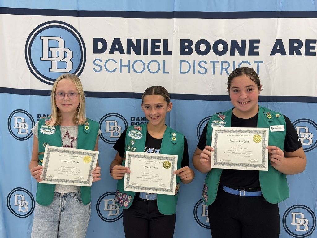Three Girl Scouts wearing their green Girl Scout vests hold their certificates and smile in front of a Daniel Boone Area School District banner