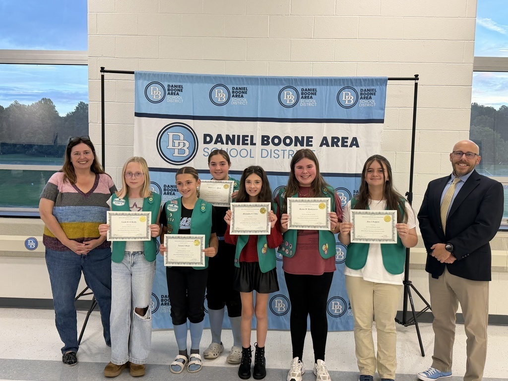 Six Girl Scouts wearing their green Girl Scout vests hold their certificates and smile in front of a Daniel Boone Area School District banner along with Superintendent Tom Voelker and School Board President Julia Olafson