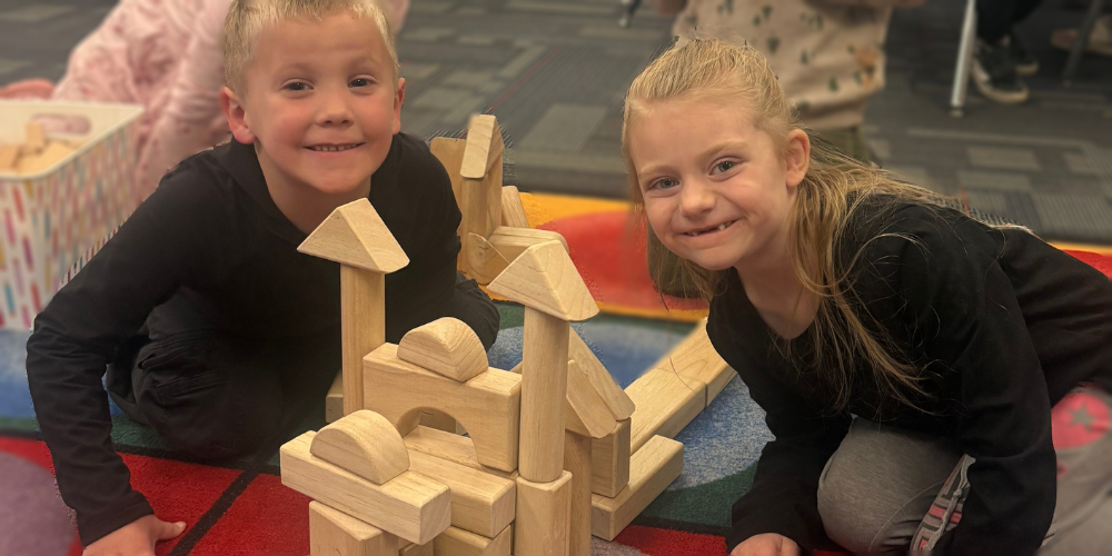 A boy and a girl in a kindergarten classroom. They are on the classroom rug with building blocks, making a castle. 