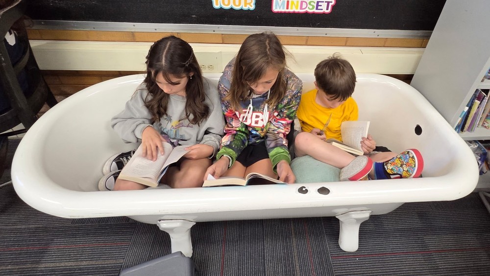 A boy and 2 girls are reading in a classroom. They are in an old bathtub with pillows.