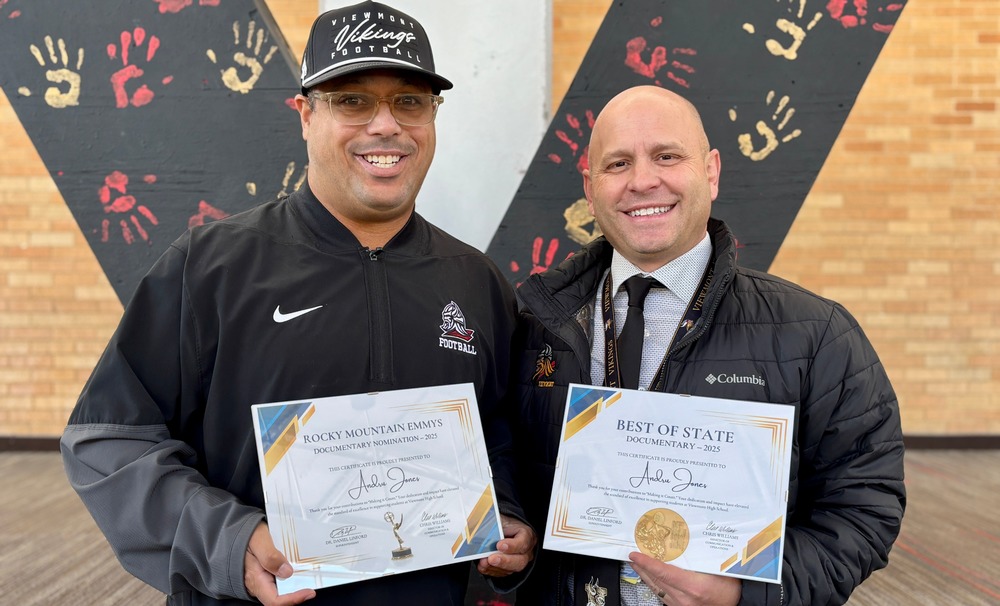 Coach Andru Jones and principal Travis Lund holding certificates in front of a big 'V' in the background with red and yellow hand prints in the Viewmont High School commons.