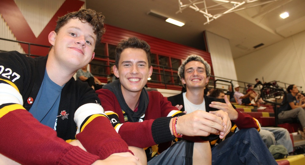 Three students sit together on gym bleachers, smiling at the camera while wearing maroon, black and gold school colors during a Viewmont High event.
