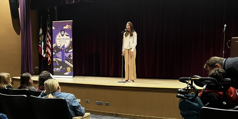 image of a student standing on a stage speaking in a microphone with spectators watching.