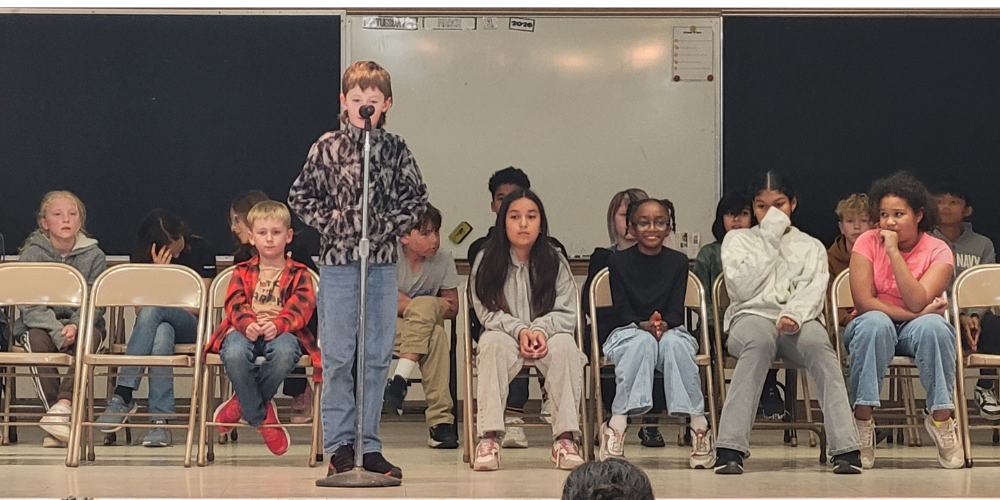 A boy is at the mic, spelling a word while the other participants sit in chairs behind him.