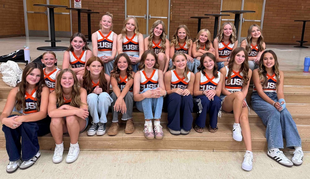 A group of 18 smiling cheerleader girls wearing their orange and blue cheer uniform tops with jeans sitting on the steps of their stage.