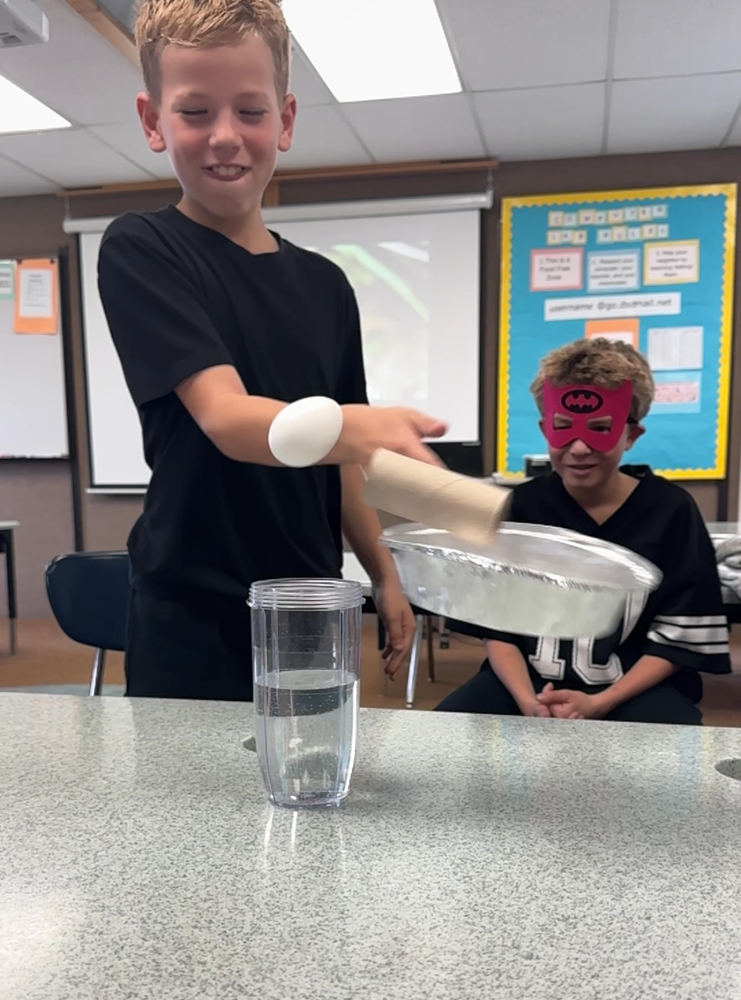 Boy knocking a pie tin with a toilet paper roll and egg off of a glass of water
