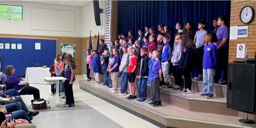 Fifth Graders are performing on our school stage during the Purple Up Day Celebration. Four students are in a line  speaking their lines in front of a microphone. Mrs. LaFrance is directing. 