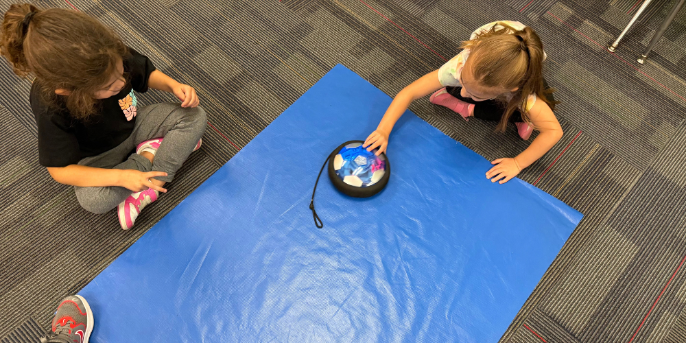 A boy and  girl at a table in the classroom pushing a circular object to learn about force