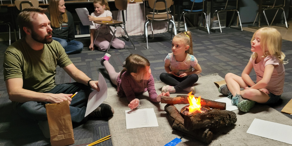 A father and 3 young firls are seated  on the floor around a fake fire in a classroom.  They are discussing words for a fill in the blank story activity. 
