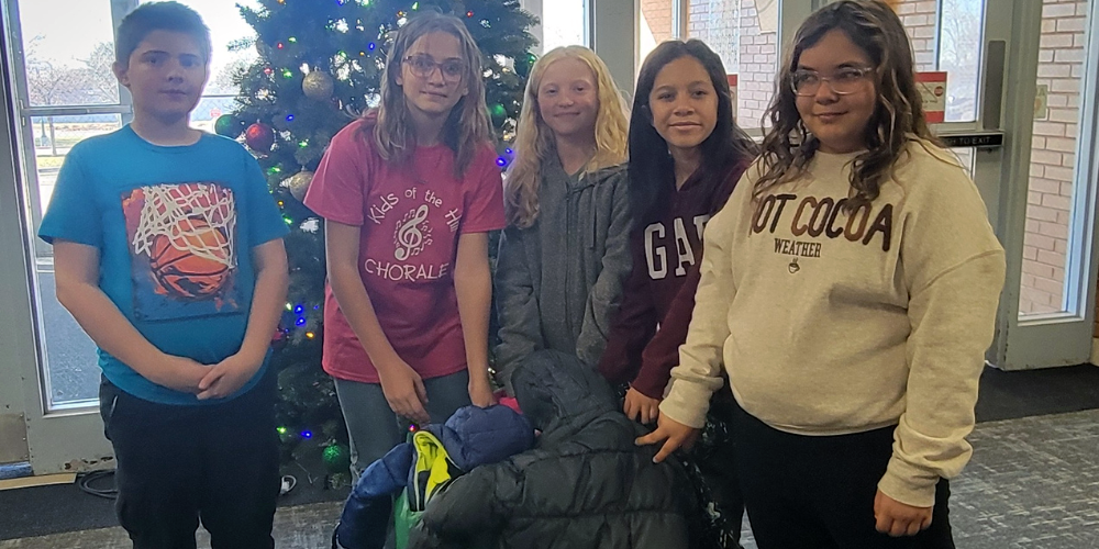 Three girls from our Anchored for Life Team are putting coats in our donation bin along with a boy and girl from our 5/6  ASC class. They are in the foyer of the school with a Christmas tree behing them. 