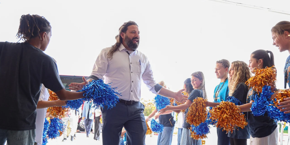 An AASA member walks through a welcome crew who is cheering and waving blue and orange pom poms at Sunburst Elementary