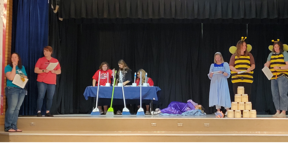 Teachers performing a play.  2 are dressed up as bees, 2 are princesses 2 students and another teacher are judges. There's a cup tower and brooms on the stage.  @ teachers are acting as children.  s.