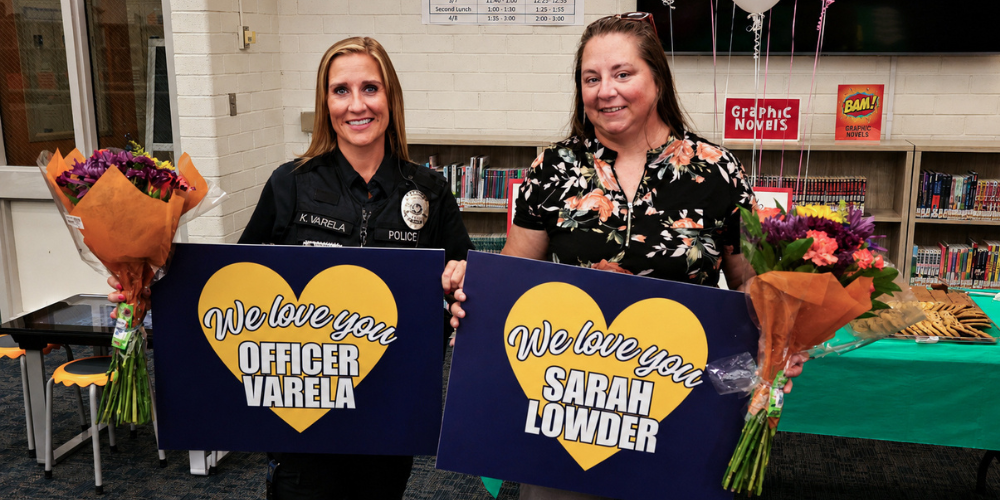 Officer Varela and Sarah Lowder smiling while holding flowers and signs that say "we love you"