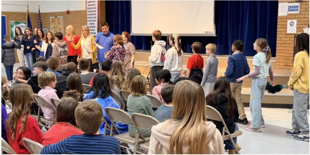Fifth graders in a line, receiving their Starbase Graduation Certificates. They are walking past Starbase teachers and classroom teachers who are congradulating them as they walk by. The audience is seated as they watch. 