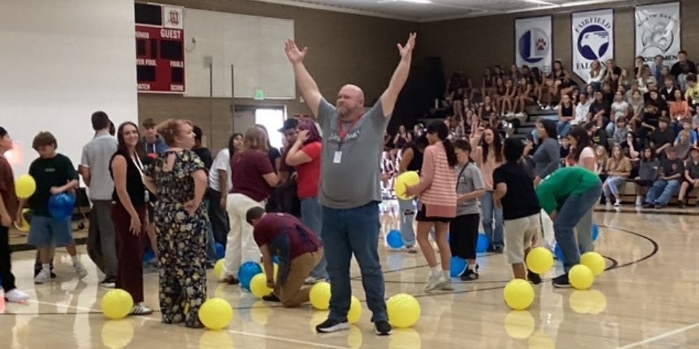A teacher with ihs arms in the air encouraging the crowd with yellow and blue balloons.