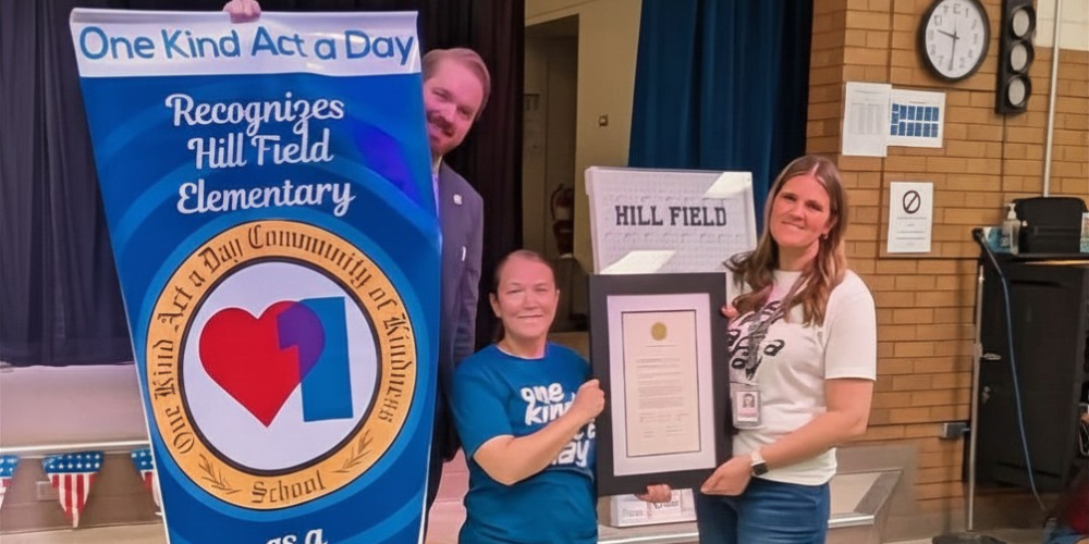 A man who is a representative of the organization One Kind Act a Day is holding a banner that says "One Kind Act a Day Recognizes Hill Field Elementary. There is a circle with an orange outer ring that says ," One Kind Act a Day Community of Kindness School." Mrs. Burk (Principal) and Mrs. Storms (PTA) are holding a plaque with a certificate of recognition.