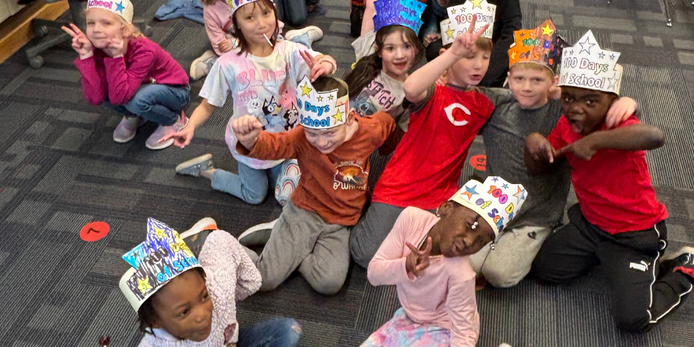 Boys and girls in Mrs. Smith's class wearing headbands that say 100 Days of School. They are sitting on the floor in a group 