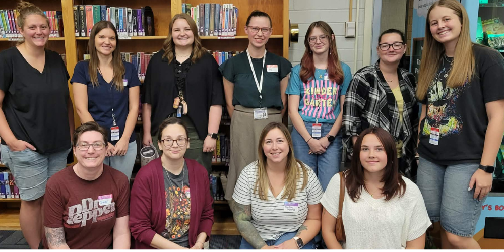 Our new staff and faculty members grouped together in front of bookshelves in the library. Seven are standing in the back row and 4 are kneeling in the front.