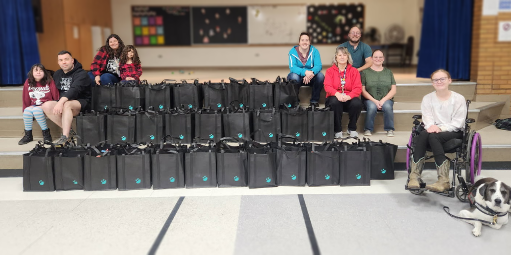 Seven adult volunteers and 2 student volunteers with 57 bags of food . They are in the school gym. 