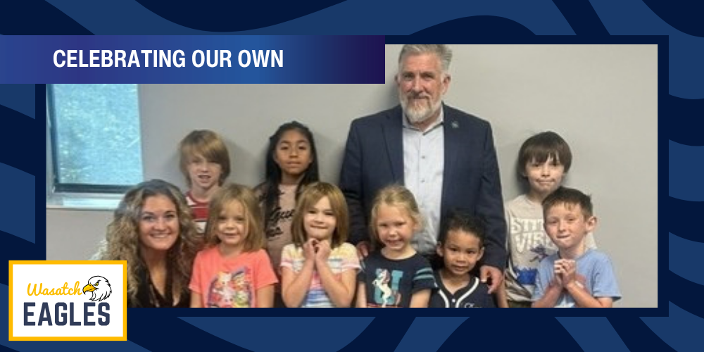 Graphic with the heading “CELEBRATING OUR OWN” at the top. A group of nine elementary students and two adults pose for a photo indoors against a light-colored wall. The image is framed with a blue Wasatch Eagles-themed border. The Wasatch Eagles logo appears in the lower-left corner with an eagle graphic and school name. The group is arranged in two rows, with students standing and seated around an adult in a blue blazer. A window is visible on the left side of the photo.