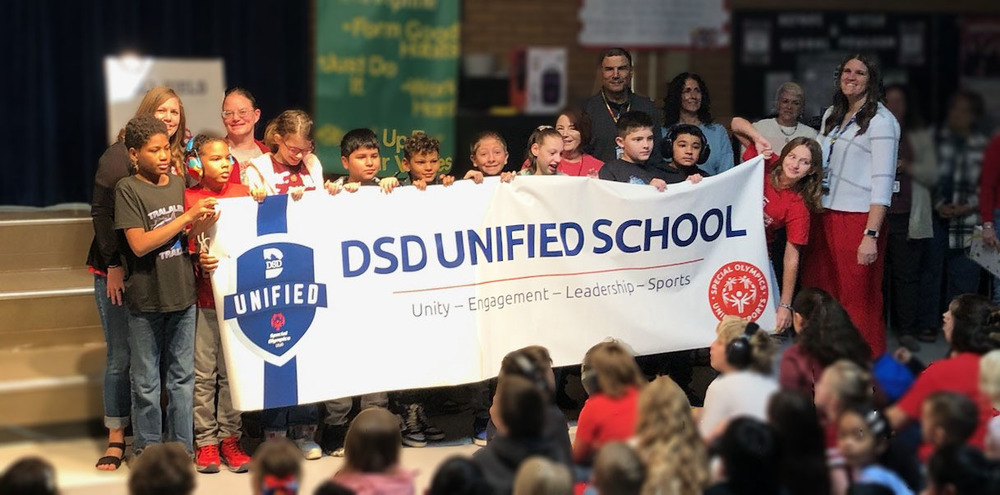 Students holding a banner that says DSD UNIFIED SCHOOL with teachers behind.